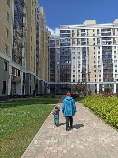 The image depicts an outdoor scene in the city of Ostafievo, Russia, on a sunny day with a clear blue sky. In the foreground, a woman wearing a blue jacket and dark pants walks away from the camera on a brick-paved pathway. Beside her, a young child in a grey jacket and green pants also walks away from the camera. The child appears to be holding something green in their right hand. To the left of the pathway is a grassy area with scattered white flowers. To the right of the pathway is a dense hedge of green leafy bushes. In the background, two large, modern apartment buildings with numerous windows and balconies dominate the scene. The buildings are constructed with light-colored brick and have contrasting dark frames around the windows and balconies. In the middle ground, between the buildings and the pathway, there is a children's playground with some play structures visible. A kite, with an orange and brown pattern, is flying in the sky between the buildings. The overall impression is a residential courtyard area.