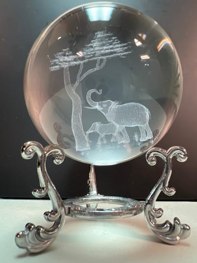 This is a close-up, eye-level, medium shot of a glass orb with an etched scene of elephants and a tree, displayed on a decorative silver stand. The orb is illuminated from behind, creating a soft glow that highlights the etched details and casts reflections on the surface. The stand is ornate, with curved metal legs that have a swirling, decorative pattern. The background is dark and out of focus, making the orb and its stand the clear subjects of the image. The surface on which the stand rests appears to be a light-colored, smooth material. There are no people or animals present in the scene other than the etched figures within the orb. The location is not provided.