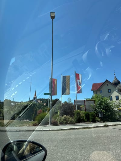 The image shows a street scene in Schonach im Schwarzwald, Germany. In the foreground, a street is visible with a sidewalk. A streetlamp stands tall, and three flags are displayed on flagpoles. One flag is red and yellow, another is black and yellow, and the third is red with a white circular emblem. A street sign that reads "Hauptstrasse" is also present. In the background, a church with a red steeple is visible on a hill, surrounded by trees and buildings. A building with a red roof and dark spire is also in the background. The sky is clear and blue. Reflections are visible on what appears to be a car windshield, including a speedometer showing "30" and parts of the car's interior like the steering wheel.