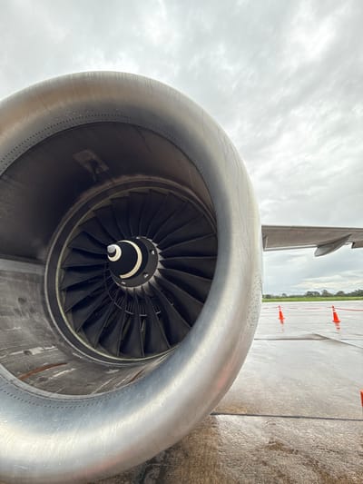 An up-close, eye-level shot shows the intake of a large aircraft engine on what appears to be an airport tarmac. The engine's metallic, silver cowling curves into the frame from the left, showing numerous rivets along its edge. Inside the cowling, a fan with many black blades is visible, converging towards a central hub which has a white and black circular pattern. The engine is positioned on a wet, concrete surface, likely a runway or taxiway, with water droplets visible on the cowling. In the background, an airplane wing extends to the right. Several orange traffic cones are spaced out on the wet tarmac, and a line of green trees and foliage can be seen in the distance under a cloudy, overcast sky. The image appears to be taken in Tarapoto, Peru, a location known for its tropical climate and frequent rainfall, which would explain the wet conditions. There are no people or animals visible in the scene.