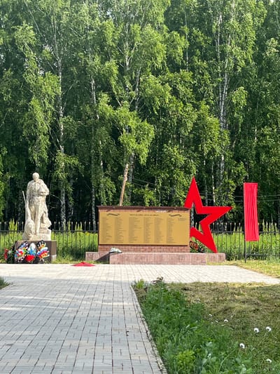 This is a ground-level, eye-level, medium shot of a war memorial located in the city of Bolshoy Gondyr, Russia. The memorial features a stone statue of a soldier holding a helmet and a flag. In front of the statue, there is a wreath of flowers. Behind the statue is a brick wall with a plaque listing names. To the right of the plaque is a large red star and a red banner. The memorial is surrounded by a metal fence. The background consists of a dense forest of birch trees with green leaves. A paved walkway leads to the memorial, and there is grass and some small white flowers in the foreground. The lighting suggests it is daytime with sunlight.