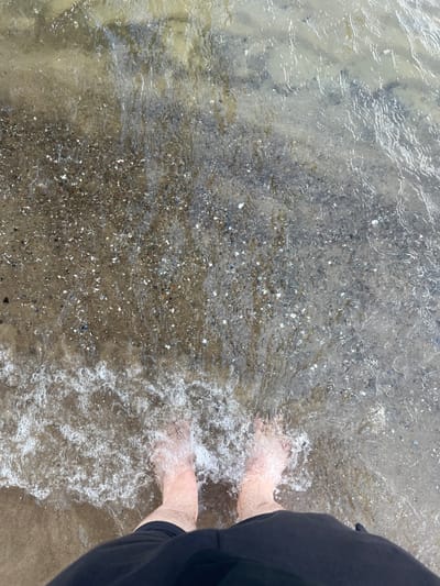 This media file captures a close-up, overhead view of a person's feet wading in shallow, clear water with a pebbly and sandy bottom. The water is slightly rippled, creating a shimmering effect on the submerged ground. Small white shells and dark pebbles are visible through the water. The person is wearing dark shorts or pants, which are visible on the right side of the frame. The scene suggests a beach or a body of water with a natural shoreline. The lighting is soft, indicating it could be daytime, possibly overcast or early morning/late afternoon. The overall mood is peaceful and relaxed, conveyed by the gentle lapping of the water around the feet. There is no visible text in the media file. The location context provided is Międzyzdroje, Poland.