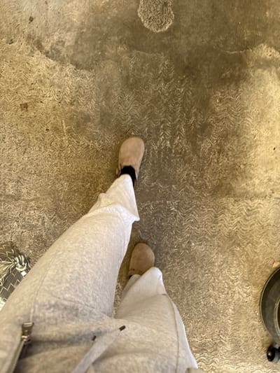 A person stands on a concrete surface wearing light gray sweatpants and beige slippers. Black socks are visible between the slippers and the sweatpants. A portion of a vacuum cleaner is in the upper right corner. The concrete surface has a rough texture.