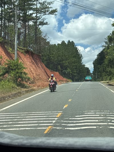 The image depicts a road scene in Thôn 6, Vietnam, characterized by a mountainous terrain with dense pine forests lining the sides of the road. The road itself is paved and appears to be a two-lane highway with white and yellow lane markings. In the foreground, a motorcyclist wearing a yellow shirt and a blue helmet is riding towards the viewer on the right side of the road. The motorcycle is dark red. In the distance, a teal-colored car is also visible, heading away from the viewer. The left side of the road features a steep, eroded embankment with reddish-brown soil, covered in sparse green vegetation and trees. The sky is partly cloudy with bright blue patches and white cumulus clouds. Utility poles and overhead wires are present along the roadside.