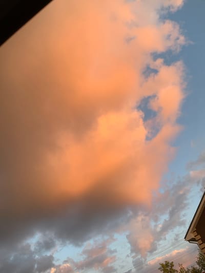 The image captures a dramatic sky with large, fluffy clouds illuminated by the warm hues of sunset or sunrise. The clouds display a gradient of colors, from a soft peachy-orange in the upper and central portions to darker, grayer tones towards the bottom and left. Patches of clear blue sky are visible peeking through the cloud cover. In the lower right corner of the image, the peak of a dark-tiled roof with brick siding is visible, suggesting the presence of a building. Some tree branches and power lines are also present in the lower part of the frame. The overall scene evokes a sense of natural beauty and the changing light conditions.