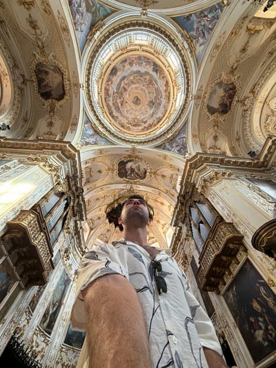The image shows a low-angle shot from inside a richly decorated church, likely in Mozzo, Italy, given the context of the provided crops. A man, seen from the chest up, looks upwards, his gaze directed towards the ornate ceiling. He is wearing a white short-sleeved shirt with a black leaf-like pattern and has sunglasses hanging from his shirt pocket. The church's interior is elaborate, featuring a large, frescoed dome, gilded architectural details, intricate moldings, and several framed paintings lining the walls. An organ console with pipes is visible on the left side of the frame, integrated into the ornate decor. The lighting appears to be natural, coming from above and the sides, highlighting the gold embellishments and painted frescoes. The perspective emphasizes the grandeur and scale of the church's interior architecture.