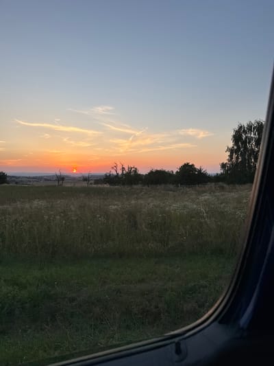The image captures a picturesque sunset over a rural landscape, as viewed from inside a vehicle. The composition is dominated by a vibrant sky transitioning from a clear, pale blue at the top to warm hues of orange and pink near the horizon. wispy clouds streak across the sky, catching the last rays of the sun.
The setting appears to be a field with tall, dry grass, suggesting it might be late summer or early autumn. Dark silhouettes of trees line the horizon, creating a natural boundary between the sky and the land. The sun, a bright orange disc, is positioned just above the tree line, casting a warm glow on the scene. In the distance, a faint outline of a town or village can be discerned. The foreground is partially obscured by the dark interior of a car, indicated by the curved edge of a window frame in the upper right and left corners of the image.
The time of day is clearly sunset, characterized by the intense colors in the sky and the low angle of the sun. The weather appears clear and calm, conducive to a beautiful sunset. The location is identified as Oberwetz, Germany, which aligns with the rural, European countryside aesthetic. There is no visible text in the image. The overall mood conveyed is one of peacefulness and natural beauty.