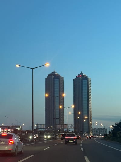 The image depicts a view from a road in Esenler, Turkey, featuring two high-rise buildings. These buildings have a similar design, with a multi-story structure and a pointed spire at the top. Each building also has a clock near the spire. The photo was taken at dusk or dawn, as indicated by the blue sky and the lit streetlights. The road appears to be a highway, with multiple lanes and cars driving on it. There are streetlights along the side of the road. The buildings and road are typical of an urban environment in Esenler.