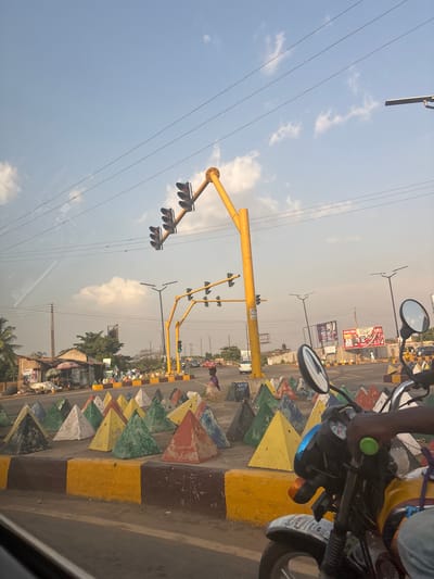 A street scene shows yellow traffic light poles with red lights against a partly cloudy sky. A row of triangular, painted concrete barriers separates lanes. A motorcycle with a mirror and green handlebar grip appears on the right. Background shows buildings and billboards. The image seems to have been taken from inside a vehicle due to the reflections on the lower portion.