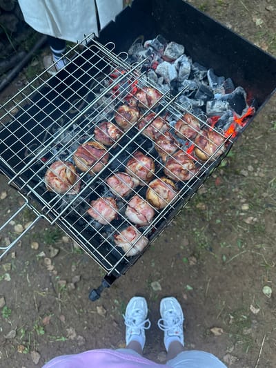 In Novaya Vozhoika, Russia, the scene is a close-up shot of a barbecue grill with food cooking. The grill is made of metal and is filled with hot coals, some of which are glowing red. On the grill, there are several pieces of food wrapped in bacon, appearing to be chicken or another type of meat. The food is positioned on a wire rack over the coals, indicating it is actively being cooked. The background shows a mix of dirt, sparse green grass, and fallen leaves, suggesting an outdoor, possibly rustic setting. In the lower part of the image, a person's feet are visible, wearing white sneakers and socks, standing on the dirt ground. The perspective is from above, looking down at the grill and the person's feet.