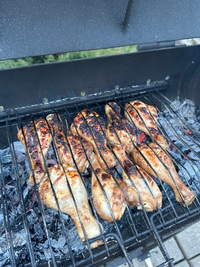The image shows chicken drumsticks being grilled on a charcoal grill. Some of the drumsticks are charred, while others are less cooked. The grill is black and sits over a bed of gray ash and embers. The location is unknown.