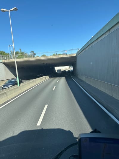 The image captures a view from a vehicle driving on a multi-lane highway. The highway appears to be an urban bypass or an elevated section, as it passes under a concrete overpass. The overpass has a pedestrian walkway with a metal railing and is supported by large concrete structures. Beyond the overpass, trees and a clear blue sky are visible.

On the highway, several vehicles are present, including cars and a van. The road surface is asphalt, marked with white lane dividers. The right side of the road is bordered by a tall, textured concrete wall, likely a sound barrier. On the left side, there is a concrete median separating lanes, with a street lamp positioned on a pole. A dark-colored car with a license plate reading "A-MI 3001" is visible in the left lane.

The image shows no people directly but implies human activity through the presence of vehicles and infrastructure. The environment is bright and sunny, indicating daytime. The overall scene is one of a typical urban highway environment.

The image includes a sign with a blue circle and a white horizontal bar, which is a traffic sign indicating a prohibition for all vehicles. Another sign with a white triangle and a red border with a blue circle inside indicates a speed limit. These signs are visible on the overpass.

While the image shows a highway in an urban setting, specific details pinpointing the location within Augsburg, Germany, are not present within the visual information provided. The license plate "A-MI" suggests the vehicle is registered in the Augsburg area, but this cannot be definitively confirmed without further context or knowledge of German license plate conventions.