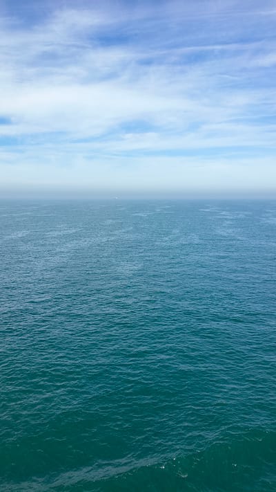 The image captures a wide, unobstructed view of the ocean stretching towards a hazy horizon under a pale blue sky. The water is a deep teal, with subtle ripples and patterns created by gentle waves. The sky above is a gradient of blue, with wispy white clouds scattered across it, particularly in the upper portion of the frame. In the far distance, a faint, light-colored object, likely a ship, is barely visible on the horizon, almost lost in the atmospheric haze. The foreground shows more detailed texture of the water, with lighter foam or spray appearing on the right side. The scene is serene and vast, conveying a sense of open space and calm. The location is identified as the city of Unknown, United Kingdom.
