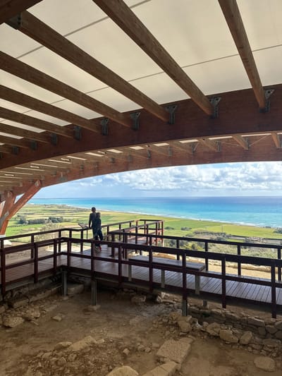 The scene depicts an outdoor archaeological viewing platform located in Piskobu Leymosun, United Kingdom. An overhead canopy, constructed from white fabric supported by robust wooden beams and metal brackets, frames the upper portion of the image. Beneath this structure, a dark-stained wooden walkway with railings extends across the midground, situated above exposed ancient stone foundations and bare earth. Two individuals, an adult and a child in dark attire, stand on this walkway, observing the panoramic coastal view. The adult is positioned near a small, dark information panel. The background reveals expansive green fields sloping towards a vivid blue-green sea under a partly cloudy sky, with a visible coastline stretching into the distance.