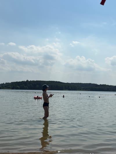 A wide shot captures a sunny day at a lake near Ozyaritska-Slabadski rural council, Belarus. In the foreground, a person wearing a bucket hat and a bikini stands knee-deep in the calm water, holding a phone. Their reflection is visible on the water's surface. Further out in the lake, a pink inflatable ring with a person inside floats, and other individuals are swimming. The background features a dense, dark green treeline bordering the lake, with a bright blue sky filled with scattered white clouds above. A small red object is visible at the very top edge of the frame.