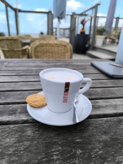 A cappuccino sits on a wooden table next to a cookie on a white saucer with a spoon. The cup is white with a brown stripe and the word "BLEND" printed in red. The background is blurred, but shows rattan chairs, a beach, and a blue sky. This scene is plausibly set in a restaurant on the beach of Bergen, Netherlands.