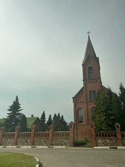 An eye-level, medium shot captures the side view of a brick church with a tall steeple, topped by a cross, against an overcast sky in Опсаўскі сельскі Савет, Belarus. The church's facade is adorned with arched windows and brickwork details. A low, decorative brick and stone wall with decorative pillars and a metal gate partially encloses the church property, separating it from the paved road and a patch of green grass in the foreground. Evergreen trees are interspersed around the church and behind the wall, with a building with a green roof visible to the left. The overall atmosphere is serene and somewhat muted due to the cloudy weather. There are no people visible in the image.