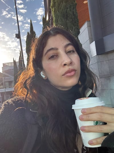 The image depicts an outdoor scene in Alcobendas, Spain, featuring a female individual. She occupies the central foreground, looking directly at the camera with a neutral expression. She has long, dark, wavy hair, wears a dark turtleneck beneath a dark, textured jacket, and has a white wireless earbud inserted in her left ear. Facial piercings include a silver septum ring and a silver labret stud below her lower lip. Her right hand is holding a white disposable Starbucks coffee cup with a white lid adorned by a small orange-red circular design. Strong sunlight originates from the left, illuminating her hair. The background includes a partially cloudy blue sky, several tall coniferous trees, and modern buildings with grey and dark facades. A dark utility pole is visible on the far left.