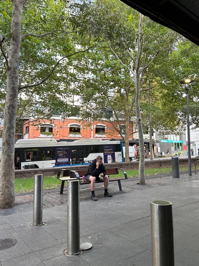 A bus stops in front of a brick building. A man sits on a bench in the foreground eating, next to metal bollards. Trees line the street. The scene is slightly tilted.