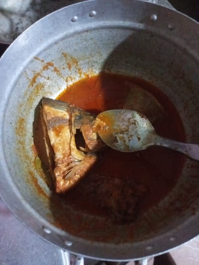 In Agbara, Nigeria, a close-up shot captures a metallic pot containing a fish head submerged in a reddish-orange sauce. A tarnished metal spoon rests within the pot, partially obscured by the fish and sauce. The pot's interior exhibits stains and specks, suggesting previous use. The lighting is dim, casting shadows and highlighting the textures of the food and cookware.
