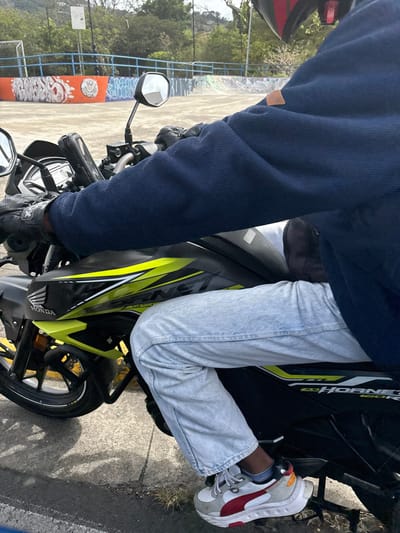 A person wearing a blue jacket, light blue jeans, and white sneakers with red accents is sitting on a black Honda motorcycle with a yellow stripe. The motorcycle is parked on the sidewalk in front of a skate park with graffiti and a metal fence. The scene is taken in San Rafael, Costa Rica, at the coordinates 9.938 latitude, -84.153 longitude.  The person is wearing a black helmet with a red visor but it is partially obscured by the jacket. The person is looking to the right of the frame.  The motorcycle is tilted slightly to the right, and the front wheel is turned to the left. The front brake lever is visible on the handlebars.  The skate park is in the background, and there are trees and buildings visible in the distance.