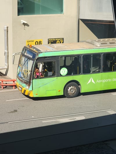 This is a media file showing a bright green airport shuttle bus, likely a Mercedes-Benz, parked outdoors on an asphalt surface. The bus is positioned at an angle, with its front facing towards the right of the frame and its side visible. The scene appears to be set at an airport or transportation hub, as indicated by signage.
On the side of the bus, the text "Aeroporto de" is visible in white, suggesting it serves an airport. A prominent circular green sign with the number "07" is also present on the side of the bus. Further down, a yellow sign with black lettering reads "PORTÃO 08 GATE" and another yellow sign with black lettering reads "PORTÃO REMOTO 07 REMOTE GATE." This suggests the bus is at a gate, possibly a remote gate. There are some visible signs of wear and tear on the bus's paint.
Inside the bus, through the windows, passengers can be seen, some appearing to be wearing colorful clothing. The lighting suggests it is daytime, likely sunny given the strong shadows cast by the bus and nearby structures. The setting includes a beige wall with a large window and a security camera mounted on it. The overall impression is that of an operational airport shuttle bus waiting to transport passengers.