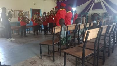 A brass band is performing in a room with rows of chairs. The musicians wear red jackets and hats, playing tubas and trumpets. In the foreground, children sit in the chairs, looking toward the band. A purple and white draped fabric is visible in the background. The room appears to be a church or community hall, with simple wooden furniture and tiled floors.