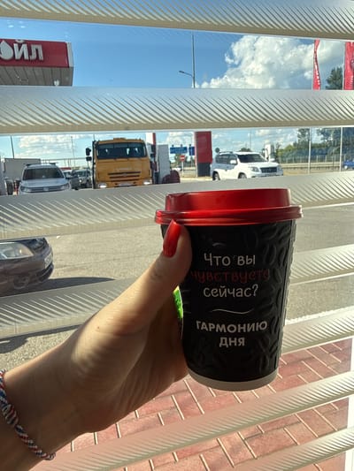 A woman's hand, adorned with a red fingernail polish and a woven bracelet, holds a black paper coffee cup with a red lid. The cup has embossed patterns and text in white. The visible text reads "Что вы чувствуете сейчас? Гармонию дня", which translates to "What do you feel now? Harmony of the day". The background appears to be a view through horizontal blinds. Outside, a street scene is visible with cars, including a yellow truck and a white SUV, and a sign with "ОЙЛ" in red letters, indicating a gas station. The location is identified as Tlustenkhabl, Russia. The lighting suggests it is daytime, with a bright sky and clouds.