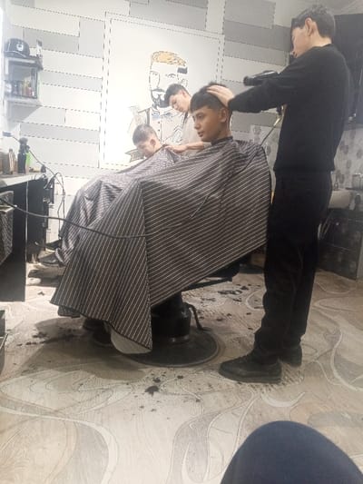 The image captures an interior barbershop scene in Laish, Uzbekistan. Four male individuals are present. In the mid-ground right, a male barber, attired in a dark long-sleeved shirt and dark trousers, stands, holding a black hairdryer aimed at the head of a seated young male client. The client, with dark hair, is covered by a black and white pinstriped barber cape. To the left, another young male client is seated in a similar dark barbershop chair, also draped with a matching cape, facing away from the viewer. Behind this second client, a third male individual, wearing a light-colored shirt, stands, appearing to attend to him. The barbershop walls on the left feature light-colored horizontal paneling, while the central wall displays a large, yellow and black stylized outline drawing of a bearded man in formal wear. A dark counter unit on the left holds assorted hair care products, with a shelf above it containing a dark cap and other objects. The floor is light-colored with a subtle grey swirling pattern and is visibly strewn with dark hair clippings. A white sink with a faucet is partially visible on the right.
