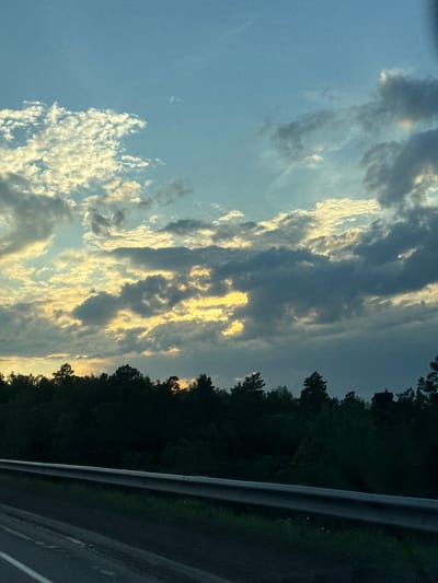 This image captures a view of the sky above a dense forest, with a road and guardrail in the foreground. The sky is a mixture of blue and gray clouds, with streaks of yellow and white light suggesting the presence of the sun, possibly at sunrise or sunset. The forest appears to be comprised of coniferous trees, silhouetted against the lighter sky. The road is paved and has a white line marking its edge. The guardrail is metallic. There are no people visible in the image. The scene suggests an outdoor environment, likely along a highway passing through a forested area. The location is identified as Malaya Kuskunka, Russia.