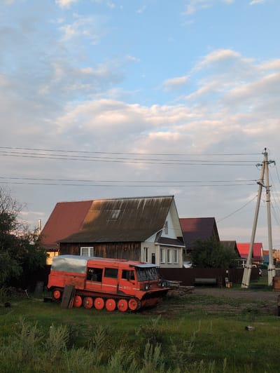 In Molochnaya Ferma, Russia, an orange tracked vehicle with a silver-topped cabin is parked on a grassy area. The vehicle has multiple sets of red wheels visible along its base. Behind the vehicle, several houses with pitched roofs can be seen, some with dark siding and others with lighter colored siding. A utility pole with power lines stretches across the upper portion of the image. The sky is partly cloudy with shades of blue and white. The foreground is filled with tall green grass and some dry vegetation. The scene appears to be an outdoor setting in a rural or semi-rural area.