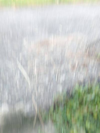 The image is blurry and appears to be a motion blur shot of a paved surface, potentially a road or sidewalk, with green vegetation on the side. Thin blades of grass or reeds are visible. The location is identified as Meunasahlancok, Indonesia, but there are no identifiable landmarks to confirm the location. No people or specific objects are discernible due to the blur.