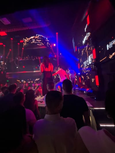 The image depicts an interior scene consistent with a nightclub or concert venue in Pamporovo, Bulgaria. The perspective is from the rear of an audience, looking towards an illuminated stage or performance area.
In the foreground, the backs of several individuals are visible, including one person wearing a white polo shirt and another in a black t-shirt. The midground shows a dense crowd of people, many with their backs to the viewer, illuminated primarily by stage lighting.
The central focus is an elevated stage area. A woman with dark, long hair, dressed in a dark, backless top or dress, stands centrally on the stage, facing away from the camera with her right arm slightly raised. To her right, a man in a light-colored shirt is partially visible, appearing to dance or interact. Other individuals are also present on the stage.
The stage is equipped with large digital screens displaying abstract, dynamic graphics with fragmented text elements, possibly "NPS," rendered in red and white. Multiple bright blue and red light beams project from various points, cutting through the darkened space and illuminating portions of the stage and crowd. The ceiling or upper structure of the venue features reflective or metallic surfaces and additional lighting fixtures. Further back, more spectators are visible on an upper level or balcony. The overall environment is dark, intensely lit by stage lights, indicating an active performance or event.