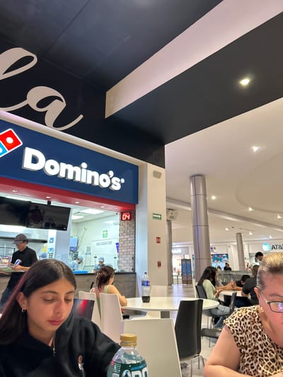 The image shows an interior dining area next to a Domino's Pizza restaurant, likely in Tampico, Mexico. The Domino's storefront is visible with its signage and order number display showing "04." Inside the restaurant, employees in uniform can be seen working.

In the foreground, two women are sitting at a table, one facing the camera with her eyes closed. A water bottle is on the table. Another woman, partially visible in the upper right, is wearing a leopard-print shirt.

In the background, several other people are sitting at tables, suggesting a public dining area in a mall or food court setting. Architectural details indicate an indoor mall environment. Logos of other establishments, including AT&T, are visible in the distance.