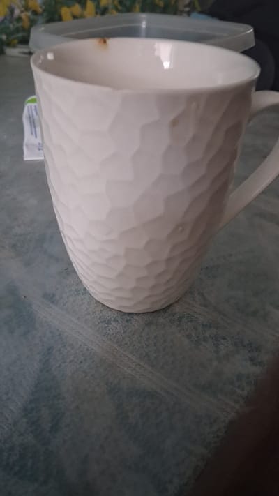 A close-up, overhead shot shows a white, textured ceramic mug with a handle. The mug is placed on a blue, patterned surface that resembles a tablecloth or placemat. A transparent plastic container with a light-colored lid is partially visible in the upper left background, along with some blurred green and yellow foliage behind it. A small, white paper packet with blue text is also to the left of the mug. There is a small brown stain on the rim of the mug. The scene appears to be indoors, likely in a kitchen or dining area.