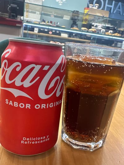 A can of Coca-Cola and a glass of Coca-Cola with ice cubes sit on a wooden table in what appears to be a cafe or bar. The can is red with white lettering, reading "Coca-Cola" in large script. Below this, smaller text reads "SABOR ORIGINAL" and "Deliciosa y Refrescante." The glass is clear and filled with a dark brown liquid with many small bubbles and ice cubes. In the background, a display case filled with pastries and a sign with blue lettering that reads "OHAH" are visible. The location is identified as La Fontsanta I Can Fatjó, Spain.