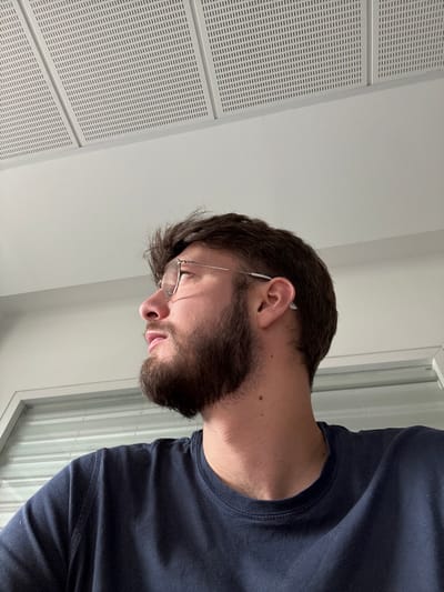 The image presents a close-up, profile view of an adult male in an indoor setting. He has short, dark brown hair and a neatly trimmed dark brown beard and mustache. He is wearing thin-framed eyeglasses and a dark blue crew-neck t-shirt. A small, white, or translucent object is visible behind his right ear. His head is turned to the left from the viewer's perspective, with his gaze directed upwards and to the left. The background features a white, suspended acoustic panel ceiling with a grid of small vertical slits across its surface. Below the ceiling is a plain white wall. In the lower left quadrant, a white horizontal frame, consistent with a window or internal opening, is partially visible, showing horizontal slatted blinds or shutters beyond. The location in the city of Lièpvre, France, cannot be ascertained from the visual content of the image.
