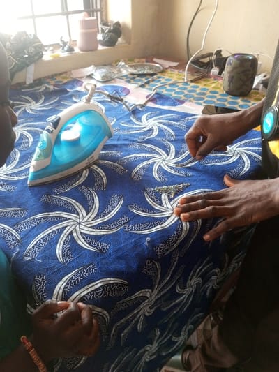 Two people are working on a blue patterned fabric. One person is using tweezers to attach embellishments while the other observes. An iron sits on the fabric near the person with the tweezers. On a table in the background are a window, a water bottle, decorative objects, embellishments, and an electronic device.
