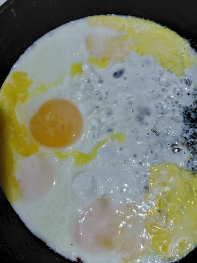 A close-up, top-down view shows fried eggs in a dark frying pan. The eggs have mostly solidified whites with a few yolks still visible, one of which is bright orange. The edges of the pan are visible as a black rim around the eggs. The scene appears to be in a kitchen in Ертіс ауылдық округі, Kazakhstan.