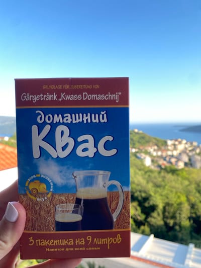 The image captures a hand holding a package of Kvass in an outdoor setting, overlooking a scenic landscape. The dominant subject is a blue and brown rectangular package, held vertically by a hand with light-colored nail polish. The package is for "Kvass Domaschnij" (Homemade Kvass), a traditional fermented drink.
The background reveals a bright, clear blue sky dominating the upper left, while the lower right shows a lush, green, hilly landscape with numerous buildings, suggesting a town or village. Beyond the land, a calm body of water is visible, likely a sea or large lake, stretching into the horizon under the bright daylight. The scene suggests a view from a balcony or terrace, characteristic of a coastal or mountainous region like Trebesin, Montenegro.
Visible text on the package, in a mix of German and Russian, includes:
- "GRUNDLAGE FÜR ZUBEREITUNG VON Gärgetränk „Kwass Domaschnij"" (German: Base for preparation of fermented drink "Kvass Homemade")
- "домашний Квас" (Russian: Homemade Kvass)
- A circular logo with a bird and text "всем знакомый вкус" (Russian: familiar taste to everyone).
- "3 пакетика на 9 литров" (Russian: 3 sachets for 9 liters)
- "Напиток для всей семьи" (Russian: Drink for the whole family)
An illustration of a pitcher filled with Kvass and a glass is also prominent on the package. The weather is clear and sunny, indicating daytime.