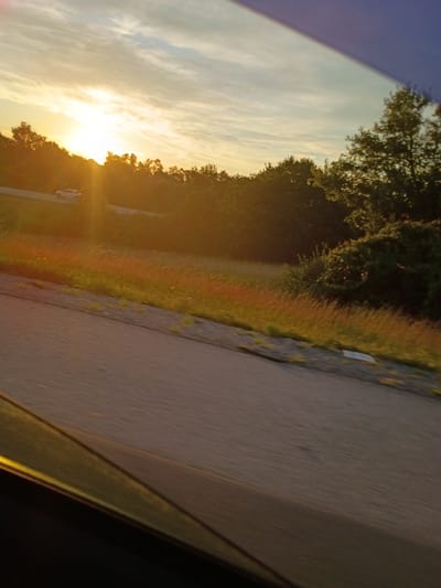 A pickup truck travels along a road through a wooded area during sunset. The sun is low on the horizon, casting a warm glow and long shadows. The sky is partly cloudy, with wispy clouds illuminated by the setting sun. The road appears to be paved and is flanked by grass and trees. A dark colored car is partially visible in the foreground, suggesting the image was taken from inside a vehicle.