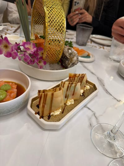 The image shows a table arrangement in a restaurant setting. Food items include a decorative plate with what appears to be stacked crepes or pancakes drizzled with syrup and topped with small dollops of cream. Adjacent is a white bowl containing salmon with green garnish and what appears to be soy sauce. Another arrangement presents oysters on ice with surrounding salmon and flowers. Part of a person's hand and arm holding a glass is visible in the background along with what looks like another plate. The location appears to be an upscale establishment. The precise location within Torreforta, Spain cannot be determined without further information.