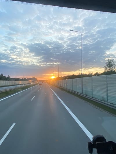 The image depicts a highway scene at sunset in Podłęże, Poland. The perspective is from inside a vehicle, looking forward. The road is paved with asphalt and has clear white lane markings. On the right side of the road, there is a tall, light-colored sound barrier. On the left side, there is a similar sound barrier and vegetation. Several cars are visible on the highway, moving in the same direction as the viewpoint. The sky is filled with dramatic clouds, tinged with the orange and yellow hues of the setting sun, which is prominently visible on the horizon, casting a bright glow and reflections on the road. Streetlights are also present along the highway. The overall impression is of a journey through a rural or suburban area during a beautiful sunset.