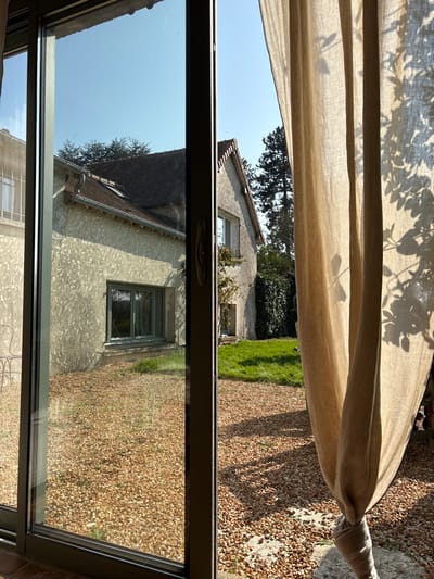 The image depicts an exterior residential scene in Bû, France, viewed from an interior perspective through a glass sliding door. The foreground features a dark-framed sliding door occupying the left two-thirds of the frame, with a tan fabric curtain partially drawn and tied back on the right, exhibiting leaf shadows from external foliage.
Immediately beyond the glass, the ground is covered predominantly by light brown gravel, transitioning to a patch of green grass on the right. Direct sunlight, originating from the left, casts distinct shadows across the gravel and grass.
In the mid-background, a two-story house with light-colored, textured walls and a dark, pitched tiled roof is central. A green-framed window is visible on its ground floor, and an additional window is present on the upper floor to the right. A gutter system runs along the left side of this house's roofline. Dense green foliage covers a section of the house's right wall, and a tall, slender evergreen tree is visible beyond it on the far right. A portion of a second building, characterized by a light-colored flat facade and a window with vertical bars, is visible as a reflection in the left pane of the sliding door. The sky above is clear and blue. No individuals are present in the scene.
