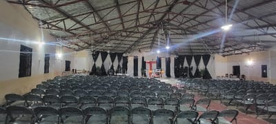 The image displays the interior of a large, empty hall or religious venue located in Itigi, Tanzania, arranged for an audience. Numerous rows of identical dark grey folding chairs with metal frames are visible, each chair bearing white alphanumeric labels (e.g., "PM 014", "PM 217") on its backrest, extending from the foreground to the rear, facing forward. A reddish floor covering or pathway delineates a central aisle. The walls are segmented, with a cream-colored lower section and a white upper section, featuring multiple dark-framed windows on both sides. The ceiling is characterized by an exposed metal truss roof structure with corrugated panels and multiple bright overhead and wall-mounted lights providing illumination.

The front of the hall, serving as the focal point, is decorated with black and white fabric drapes; diamond-patterned drapes are on the far sides, and vertically striped drapes are centrally placed. A prominent red cross stands elevated on a platform in the center. Directly below the cross, a white-covered table, resembling an altar, is adorned with floral arrangements and flanked by green potted plants. To the right of this central setup, an additional table with chairs is visible. No individuals are present, and no actions or interactions are occurring within the scene.
