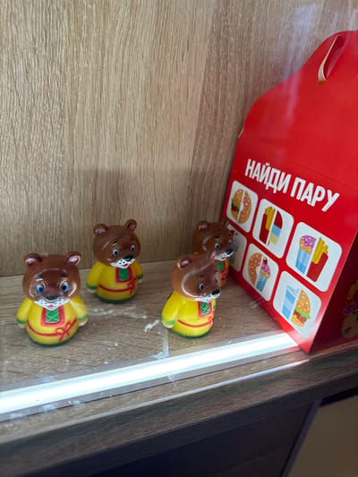 The media file captures an indoor scene, likely in Krilovo, Russia, featuring a red cardboard box and three small toy bears arranged on a light-colored wooden surface. The box, which appears to be a fast-food or kids' meal container, stands upright and displays prominent white Cyrillic text that reads "НАЙДИ ПАРУ" (NAYDI PARU), translating to "FIND A PAIR."

Below the text, the box features six square illustrations arranged in two rows. These graphics depict various fast-food items, including french fries with different toppings (one purple, one red), hamburgers, and donuts, suggesting a matching or memory game printed on the packaging.

In front of the box, three identical toy bears are lined up on the wooden surface. Each bear has a glossy brown head with blue eyes and a small snout. Their bodies are yellow, intricately decorated with red and green patterns, giving them a distinct, possibly traditional, aesthetic. The bears are uniformly positioned, facing forward towards the viewer.

The scene is illuminated by a bright, vertical strip of artificial light visible along the right edge of the frame, indicating an indoor setting. The wooden surface displays a clear grain texture, consistent with a table or shelf. No people are present, and no specific activity is taking place, but the items suggest the aftermath of a meal or a play session. The time of day is not discernible due to the indoor lighting.