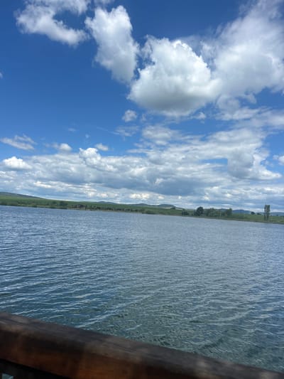 The image depicts a serene lakeside scene under a bright blue sky dotted with fluffy white clouds. The foreground features a dark wooden railing, suggesting a viewpoint from a pier or balcony overlooking the water. The lake itself is calm, with gentle ripples reflecting the sky. In the distance, a green, rolling landscape stretches across the horizon, dotted with trees and some buildings, possibly indicating a rural or semi-rural area. The overall impression is one of tranquility and natural beauty. The location is identified as the city of Begunovtsi, Bulgaria.