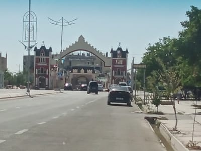 The image depicts a street scene in the city of Romitan, Uzbekistan, on a clear, sunny day. A wide, paved road runs through the center of the frame, with white dashed lines marking the lanes. Several vehicles are visible on the road, including a black sedan in the foreground, a dark SUV, and other cars further in the distance.
On the left side of the road, there is a distinctive architectural structure resembling a gateway or entrance, with a large archway and decorative towers that appear to be in a castle-like style. The buildings flanking the archway are brightly colored, with red and white facades. A tall, thin pole with an unusual decorative top is also present on the left.
On the right side of the road, there are trees with dense green foliage, a sidewalk, and a sign that reads "NOTARIUS". A person in a light-colored garment is walking on the sidewalk in the distance. The environment suggests a developed urban area with pedestrian walkways and landscaping.