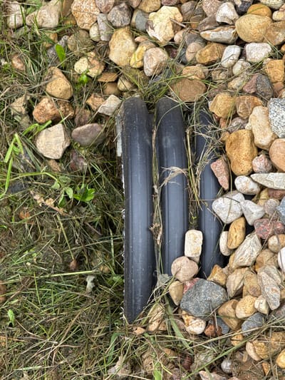 A close-up, eye-level, outdoor shot shows three black plastic pipes lying parallel on the ground. They are nestled amongst a scattering of light brown and gray pebbles and blades of green and dry grass. Some of the pebbles are roughly the size of a thumbnail, while others are larger, up to a couple of inches in diameter. The grass is a mix of green and brown, with some strands appearing dry and brittle. The pipes have a ribbed texture and appear to be slightly worn. The overall scene suggests an uncultivated or natural outdoor environment, possibly a garden or yard.