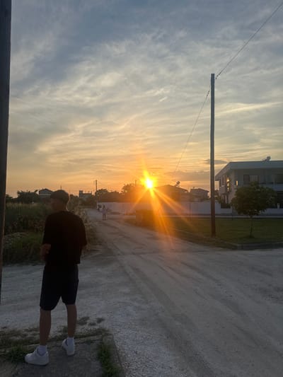 The image depicts a rural or suburban street scene in Chaidefto, Greece, during sunset. A young person, viewed from behind, stands on the left side of a dirt road wearing a black t-shirt, dark shorts, white socks, and white sneakers. They are looking towards the right of the frame, where the sun is setting with strong lens flares. In the background, several residential buildings are visible, along with scattered trees and utility poles. Two other figures, appearing to be children, are walking further down the road. The sky is filled with wispy clouds, and the overall lighting suggests the golden hour. The environment appears to be an undeveloped or semi-developed area with a mix of vegetation and constructed elements.